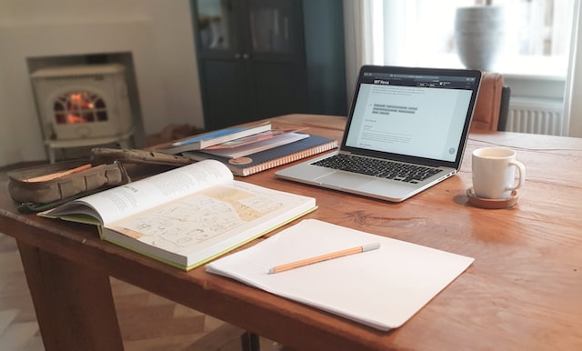 Brown wooden table set up as a desk with multiple books, a laptop, a cup of coffee, and a notebook and pen